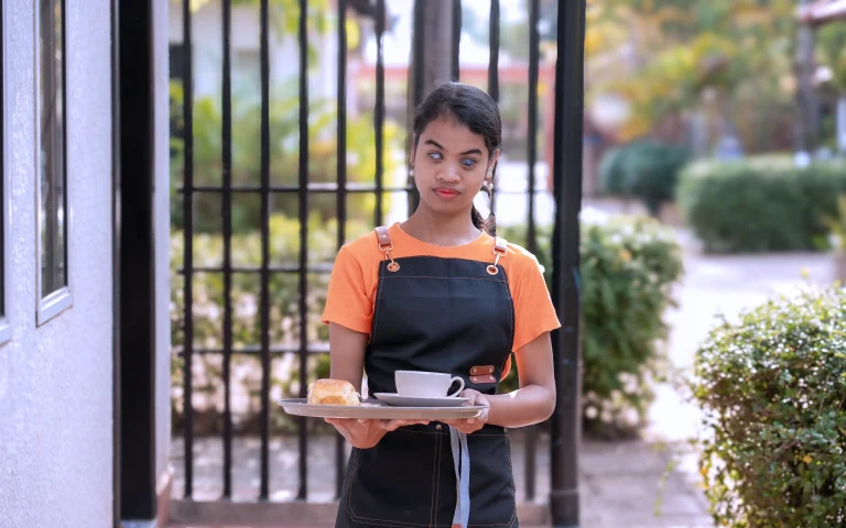 Learner with visual impairment serving coffee and pastry to a guest during café training at the DYP Learning Center in Siem Reap.